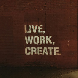 Photo by Jon Tyson on Unsplash of a white stencil graffiti on a dark red brick wall reading live, work, create in bold capital letters.