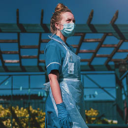 Photo by Luke Jones on Unsplash of a female healthcare worker wearing a blue uniform, face mask, gloves, and a clear plastic apron, standing outdoors under a wooden pergola on a sunny day.