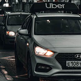 Photo by Viktor Avdeev on Unsplash of a white Hyundai car with an Uber rooftop sign and headlights on, driving in city traffic with buildings and streetlights in the background at dusk.